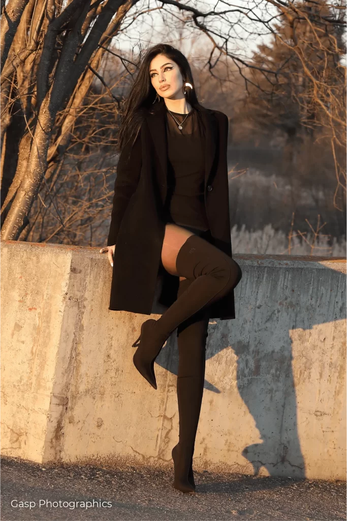 A woman in black thigh high boots leaning against a wall.